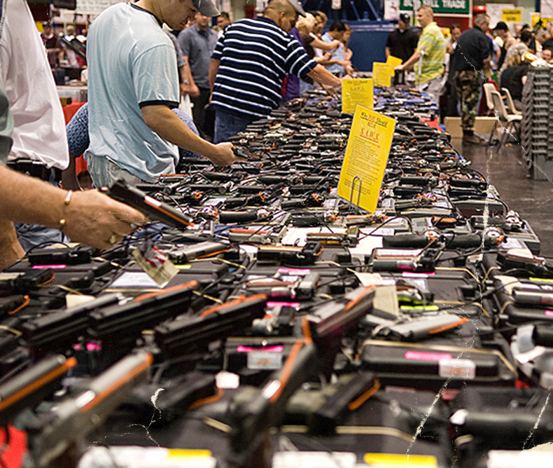 Gun show showcasing a long table with a variety of handguns with people checking them out.