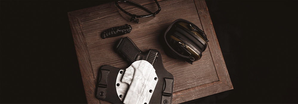 Gun safety glasses, ear protection, a pocket knife and a handgun inside a gun holster from Vedder Holsters on top of a piece of wood.